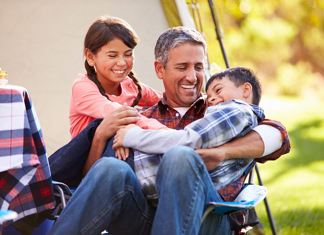 About Our Agency - Closeup Portrait of a Happy Mature Man Sitting Outside a Tent Having Fun Camping with his Two Kids on a Sunny Day