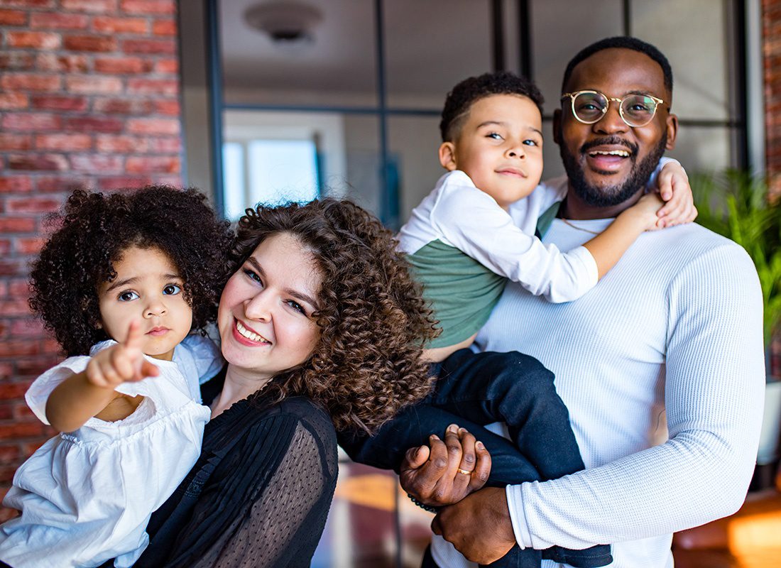 Personal Insurance - Portrait of Smiling Parents Holding Up Their Young Daughter and Son as They Stand In Front of Their Red Brick Home